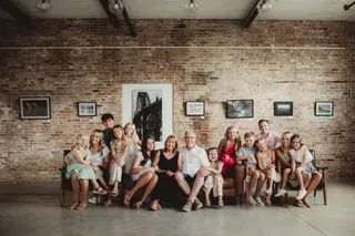 A large group sits together on a long couch in a brick-walled room.