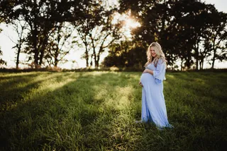An expecting person in a light blue dress stands in an open grassy field beneath trees.