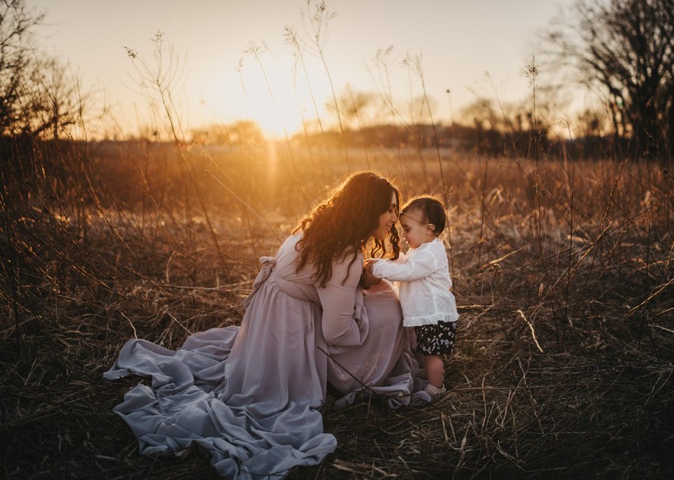 A mother in a lilac dress kneels and touches foreheads with her toddler in a sunlit field.