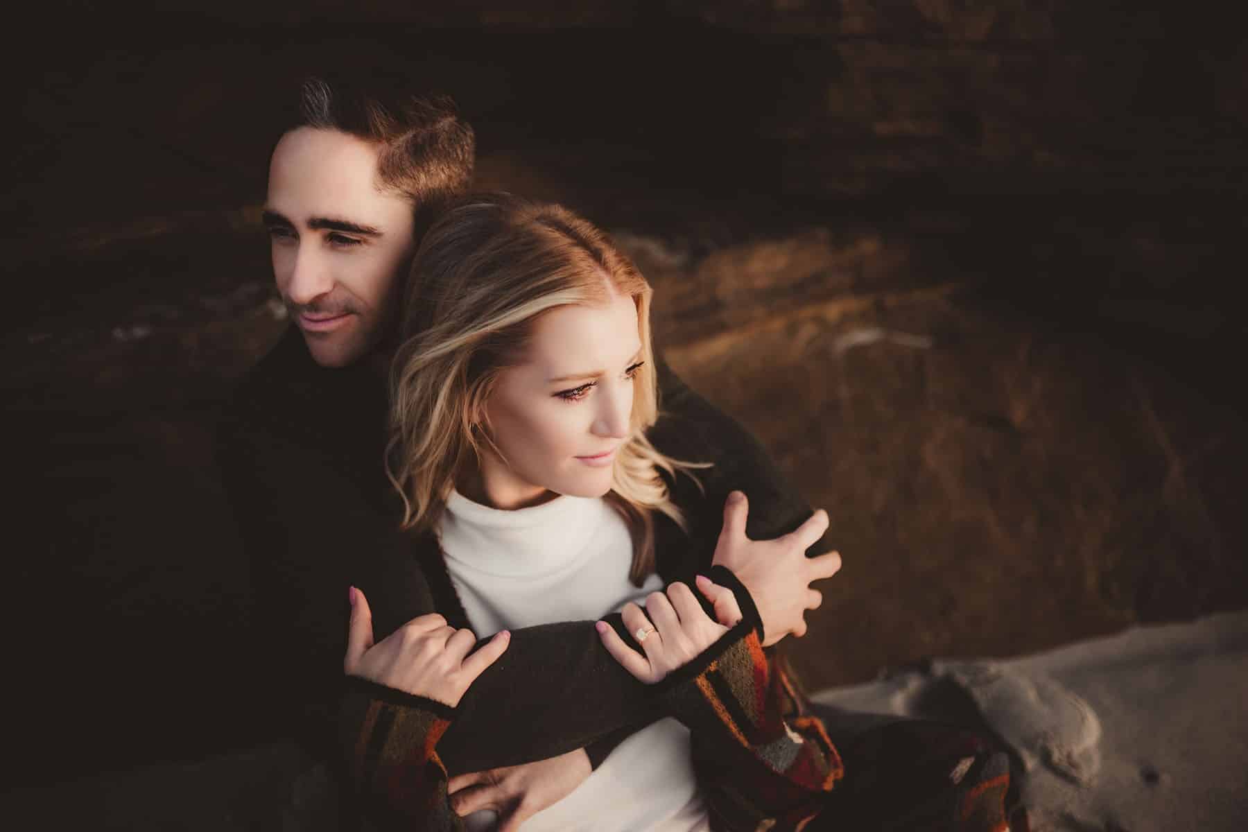 A couple embracing warmly, the man behind the woman, both facing a rocky backdrop under soft sunset lighting.