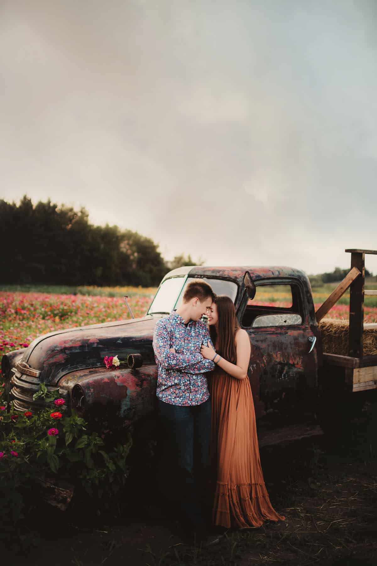 A young couple embracing beside an old rustic truck surrounded by a field of flowers at dusk.