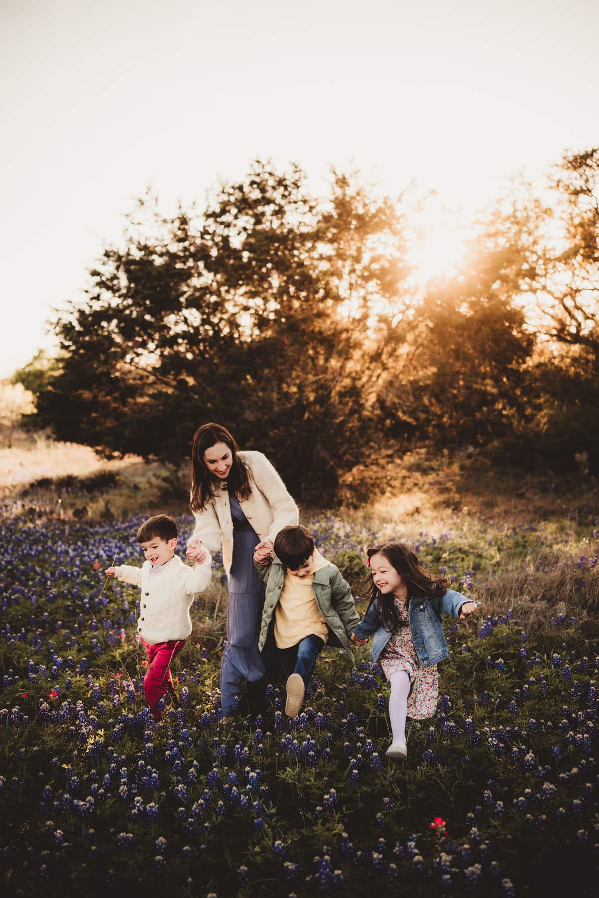 A woman and three young children happily running through a field of bluebonnets at sunset, with trees and warm sunlight in the background.