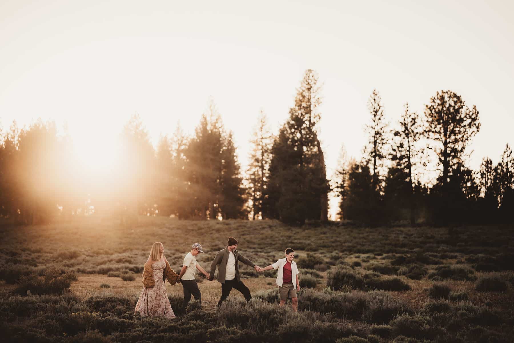 A family of five holding hands and walking through a field with a forest backdrop at sunset