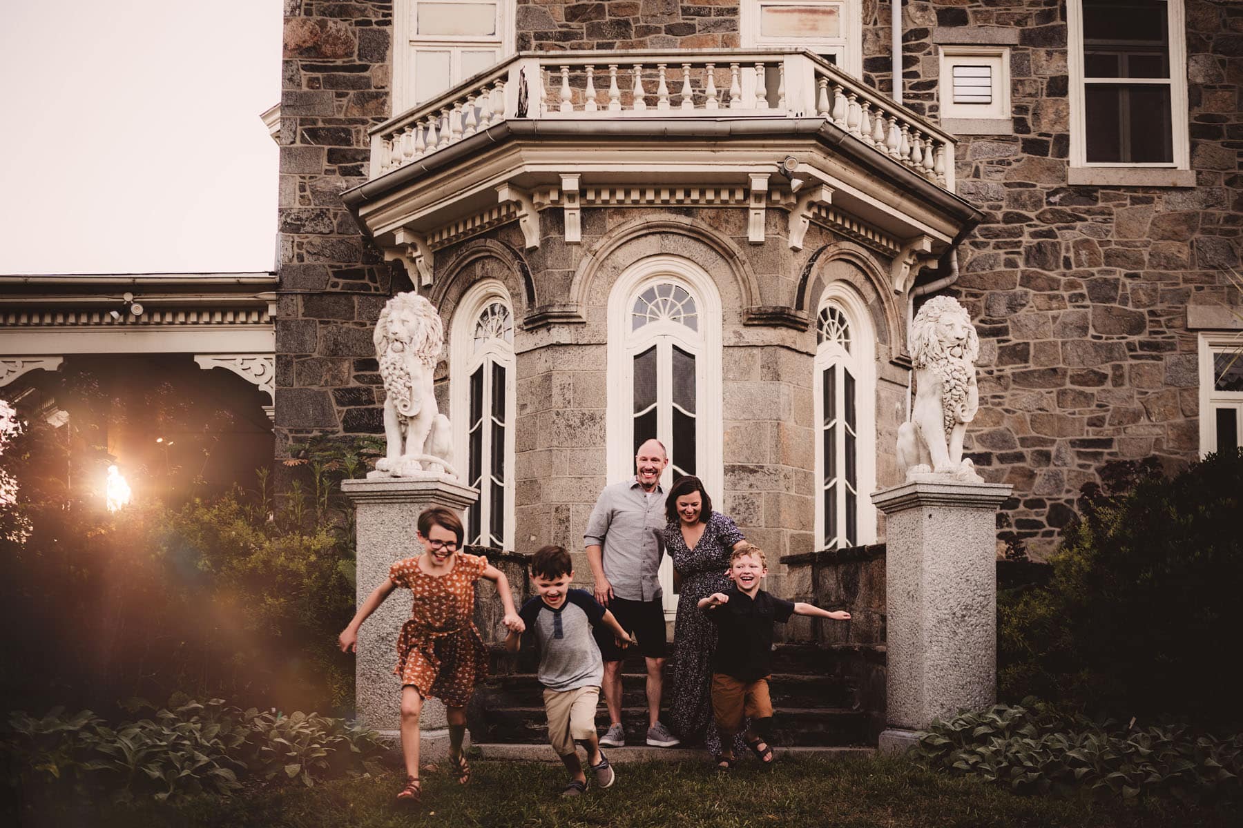 A family of five smiles and plays in front of an elegant old mansion with stone lion statues at sunset.