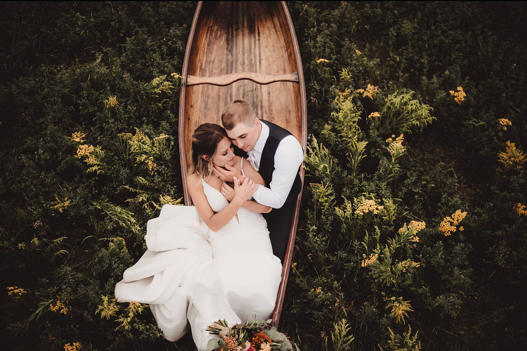 Aerial view of a newlywed couple embracing in a wooden canoe surrounded by lush greenery and wildflowers.