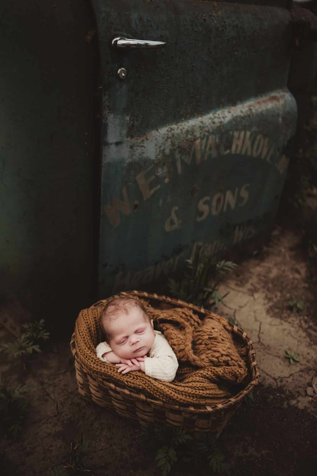 A newborn baby sleeps peacefully in a woven basket beside a weathered metal container with faded text in a natural outdoor setting.
