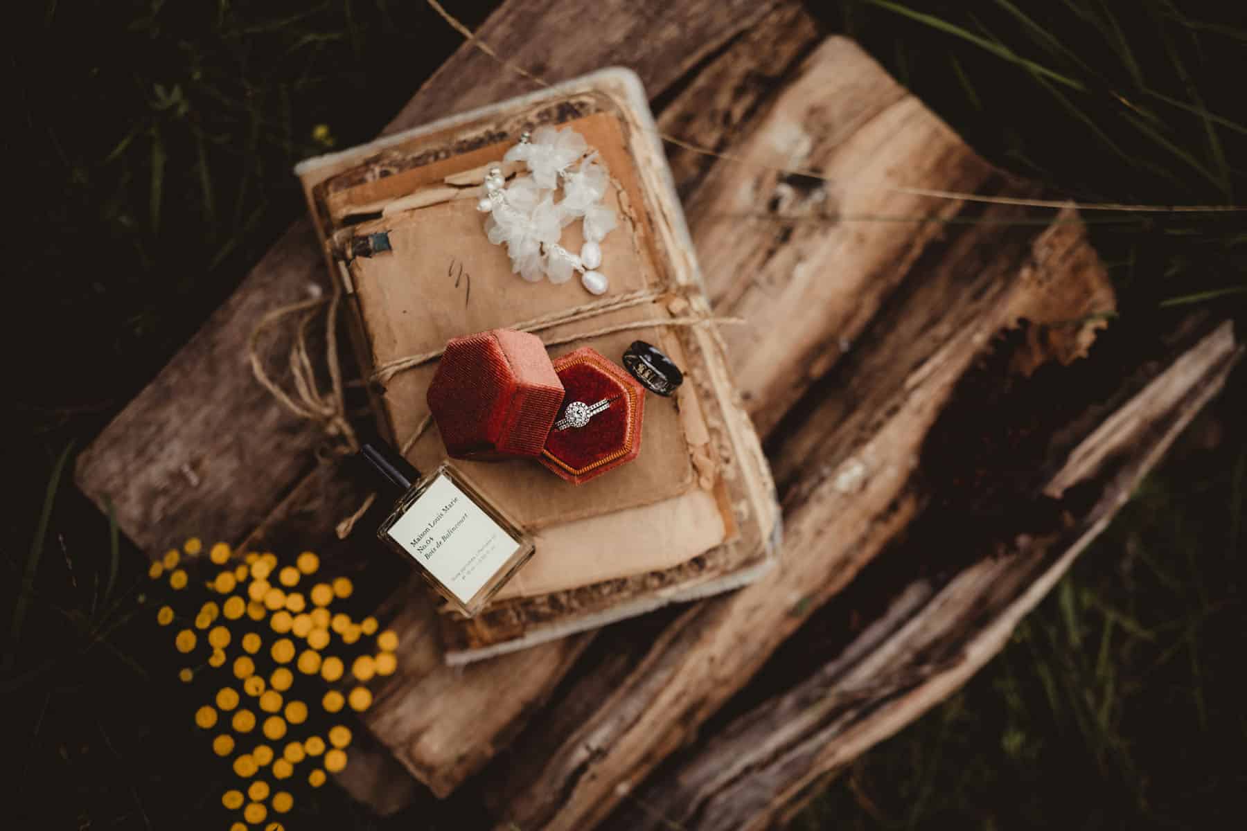 A vintage engagement ring inside a red box placed on an aged book surrounded by tansy flowers and a bottle of essential oil in a rustic outdoor setting.