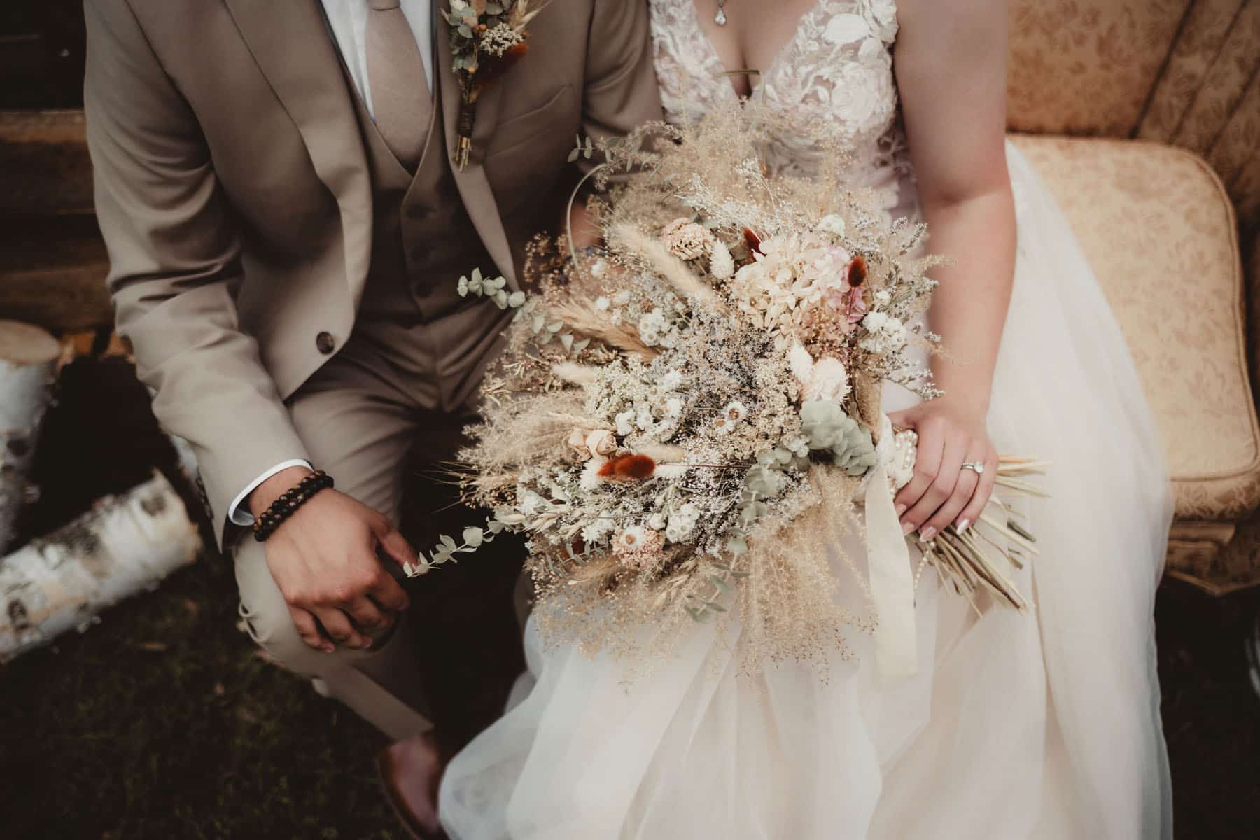 Close-up of a bride and groom holding a lavish, textured floral bouquet during their wedding, with focus on their intertwined hands and the bouquet.