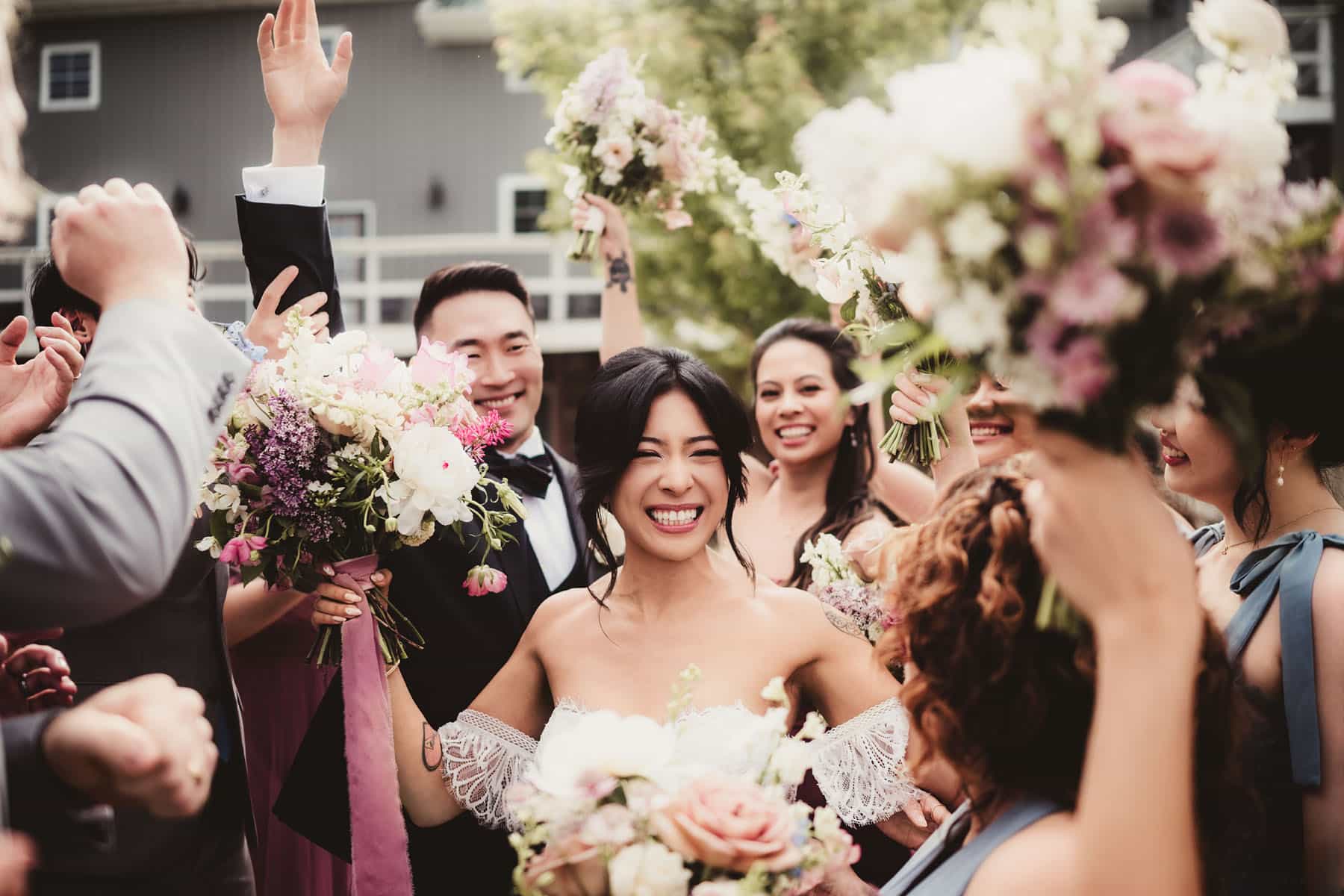 A bride at Harvest Moon Pond with a bright smile, surrounded by friends raising flowers in celebration at an outdoor wedding.