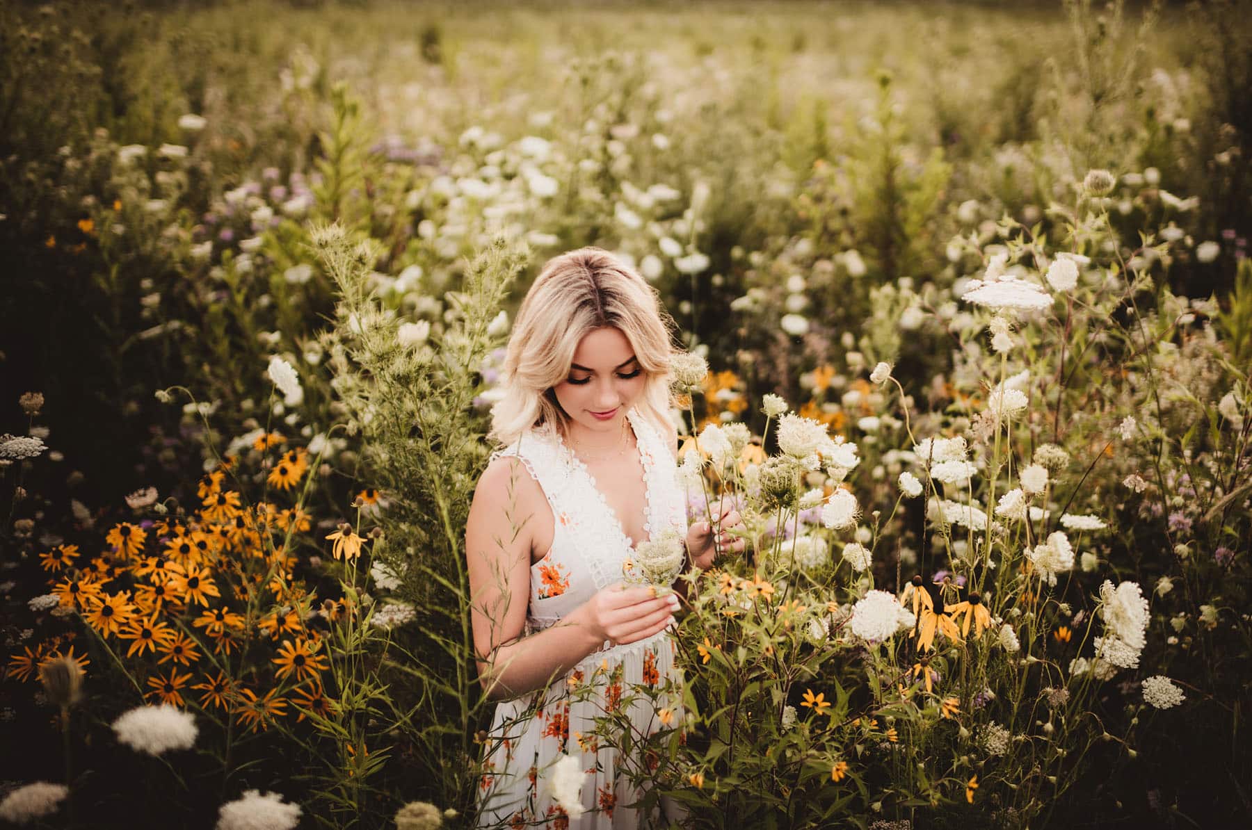 A woman in a white floral dress admiring and touching wildflowers in a lush meadow