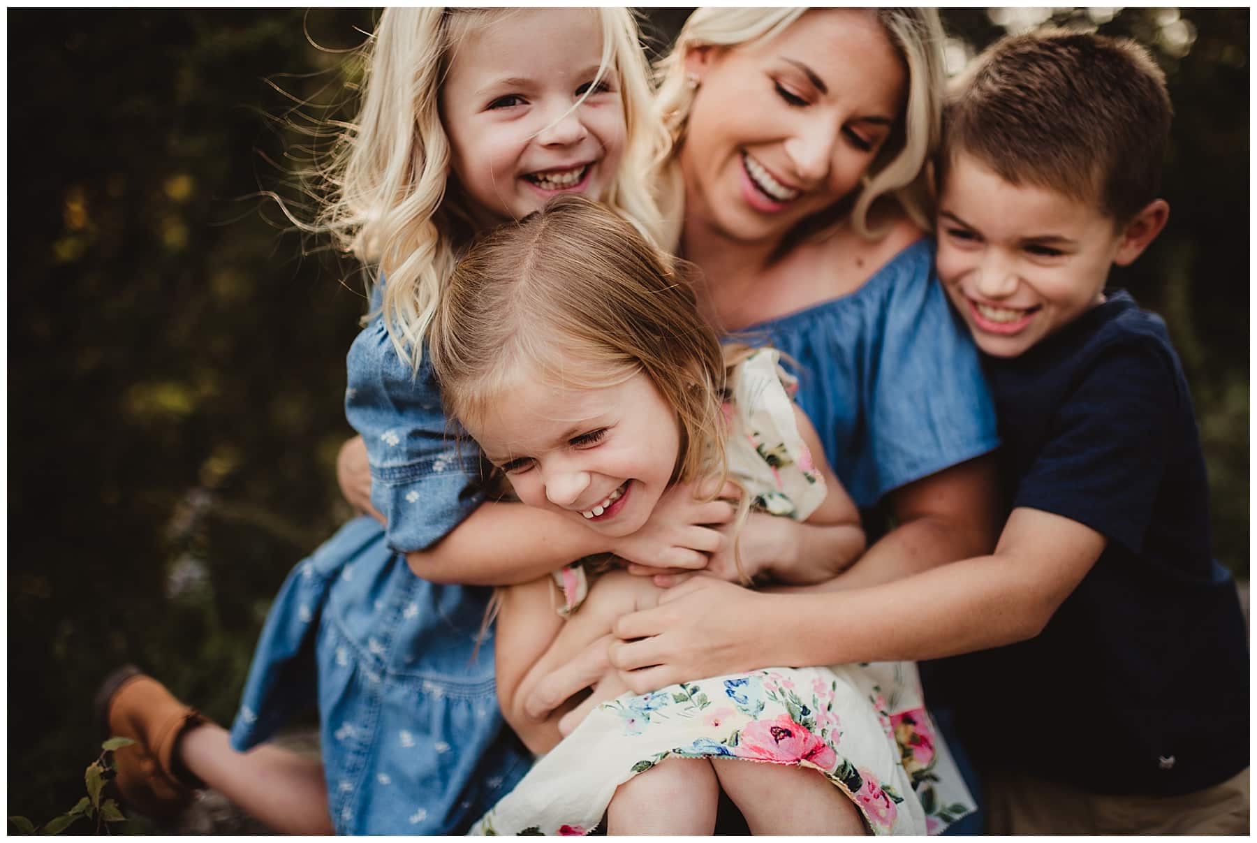 Mom laughing as her three young children pile on and hug her during outdoor family portraits