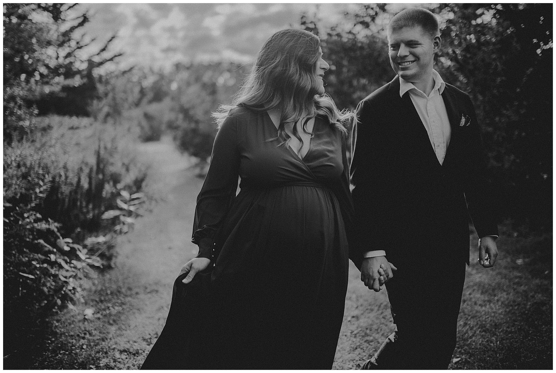 An expecting couple holding hands and smiling at each other on a wooded path, in black and white.