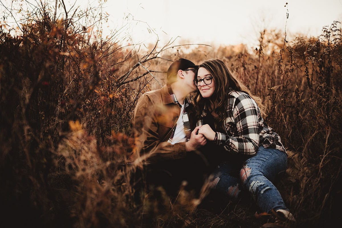 A young couple sitting close together in a field of tall autumn grasses, with the man gently kissing the woman's cheek as they hold hands.