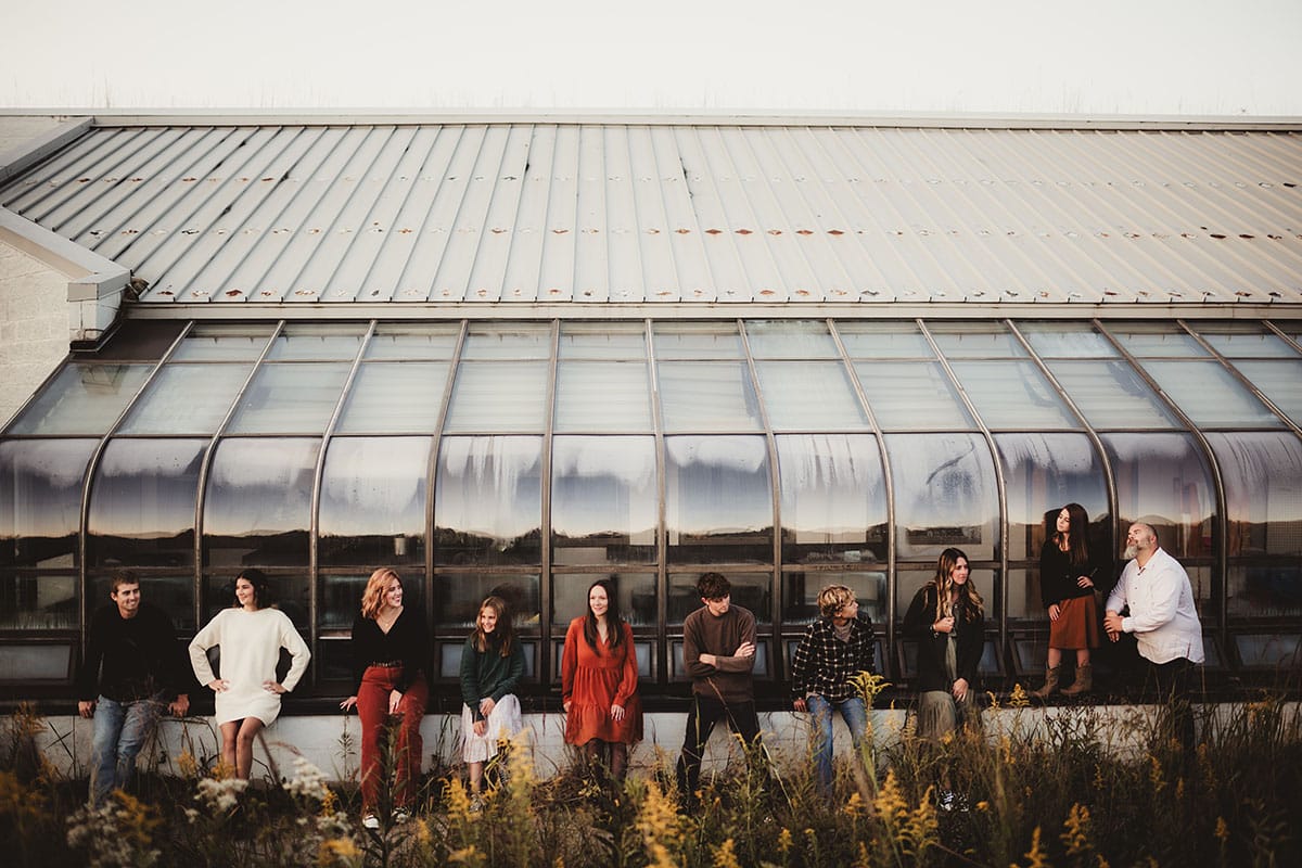 A group of people sitting and standing against a large greenhouse window, each with distinct poses, surrounded by autumn foliage.