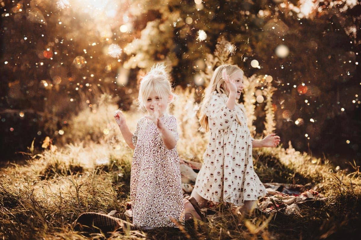 Two young girls in floral dresses playfully surrounded by dandelion seeds in a sunlit field, expressing joy and wonder.