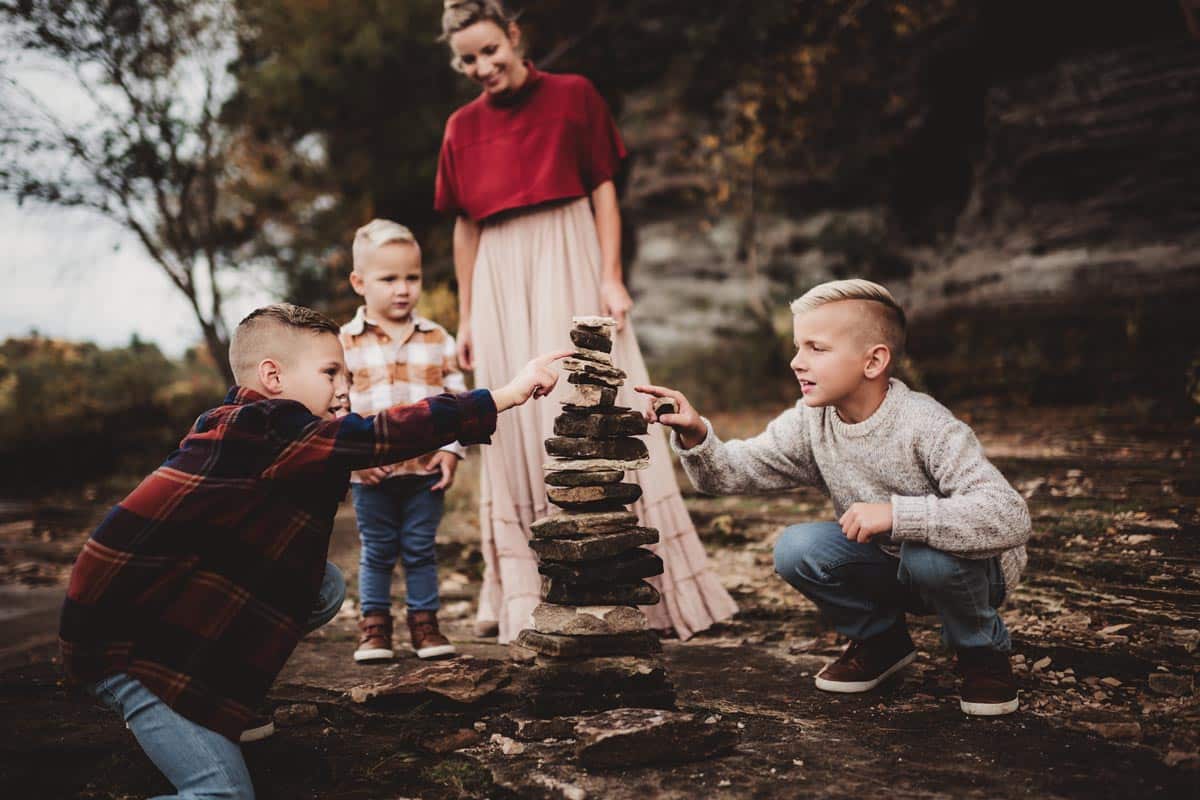 Three young boys building a stone tower outdoors, with their mother watching in the background, surrounded by autumn foliage.