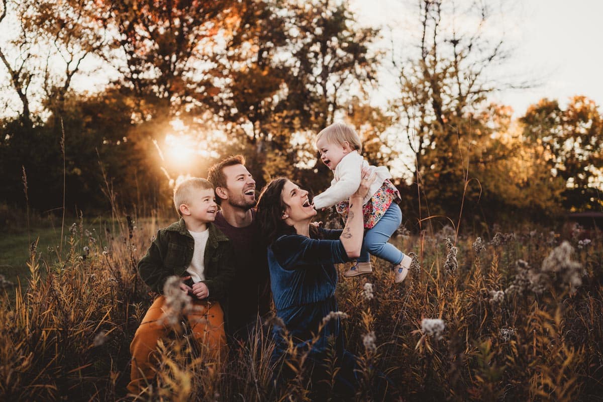 Family Portrait in Autumn Sunset Field A happy family of four enjoying a beautiful sunset in a field, with autumn colors in the background. The mother is holding up a laughing toddler while the father and young son smile warmly.