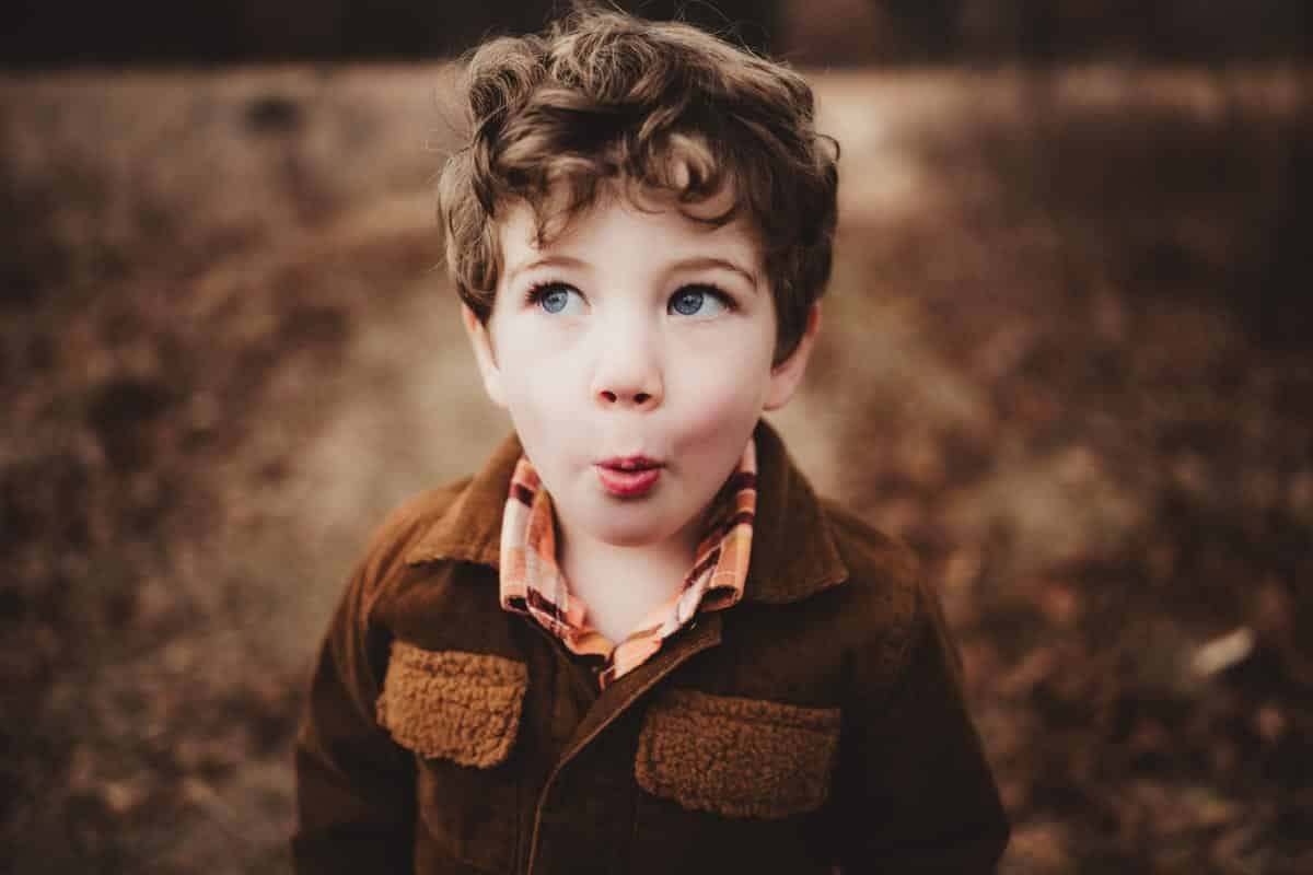 A young boy with curly brown hair and bright blue eyes makes a playful face while standing on a leaf-covered path wearing a brown jacket and plaid shirt.