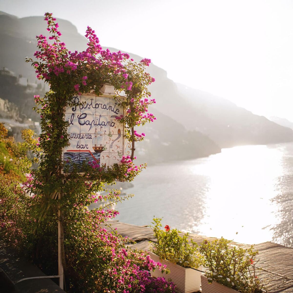 A vibrant restaurant sign surrounded by blooming flowers overlooking a serene coastal view at sunset, showcasing techniques about an online photography workshop.
