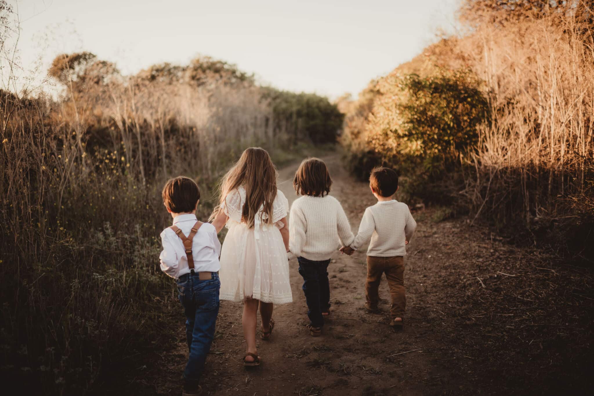 Four children walking hand in hand along a narrow path through an overgrown field during golden hour.