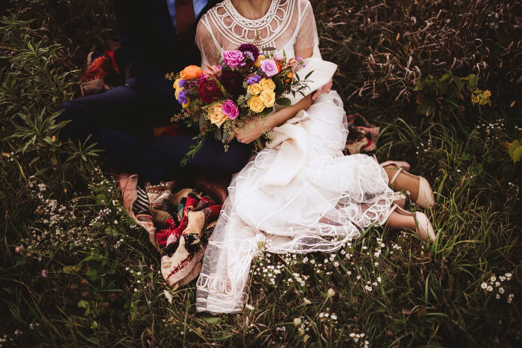 Bride and groom sitting in a lush meadow, the bride holding a vibrant bouquet of flowers