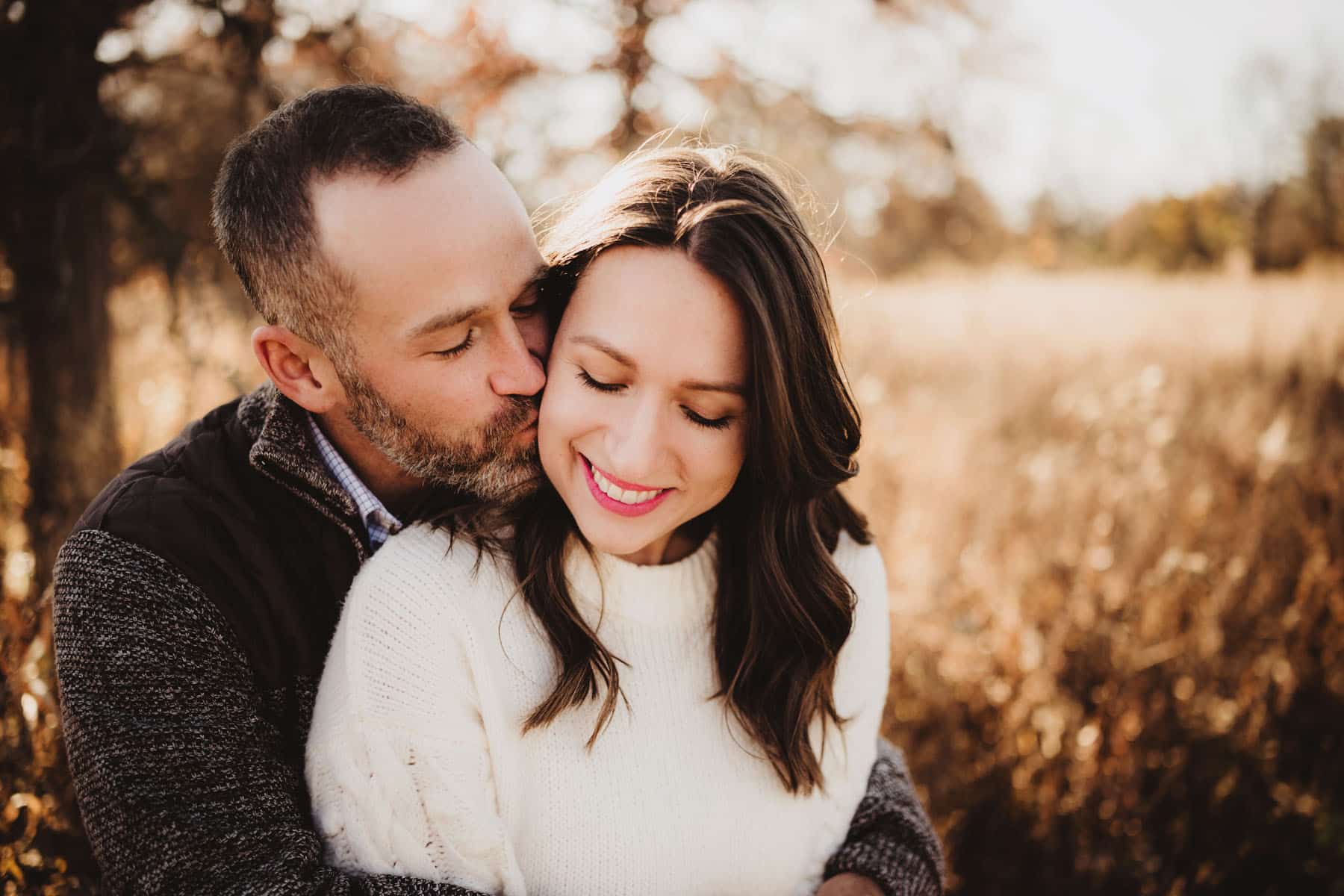 Man kissing woman on the cheek in a sunny autumn field, both smiling gently