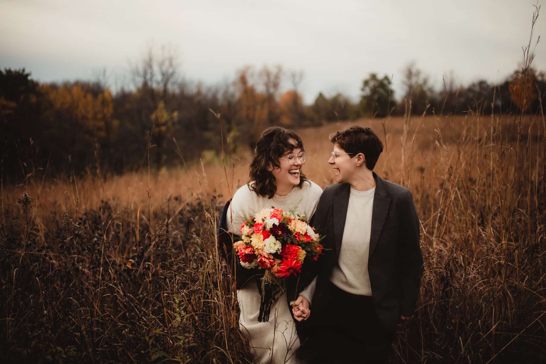 Two women laughing joyfully in a field with one holding a vibrant bouquet of flowers, illustrating an autumn wedding scene.