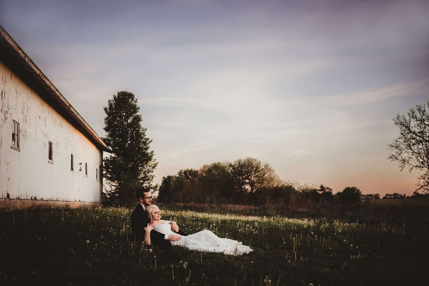 A couple lying on a grass field next to a white barn under a beautiful sunset