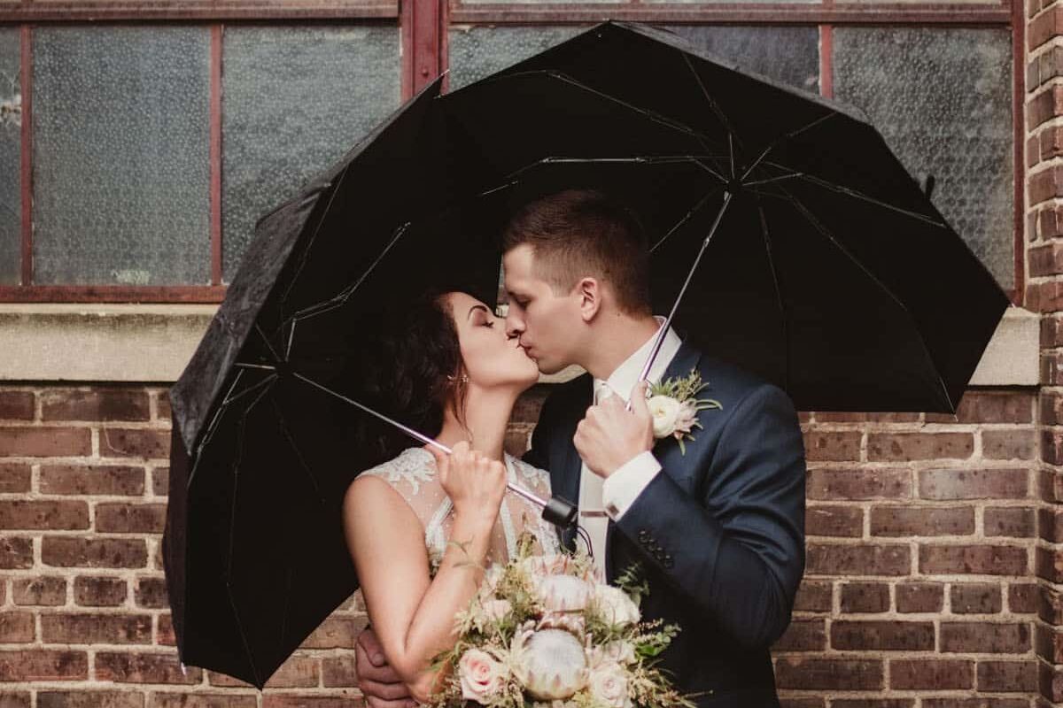 A bride and groom sharing a romantic kiss under a black umbrella, standing in front of a vintage brick building with a large window.