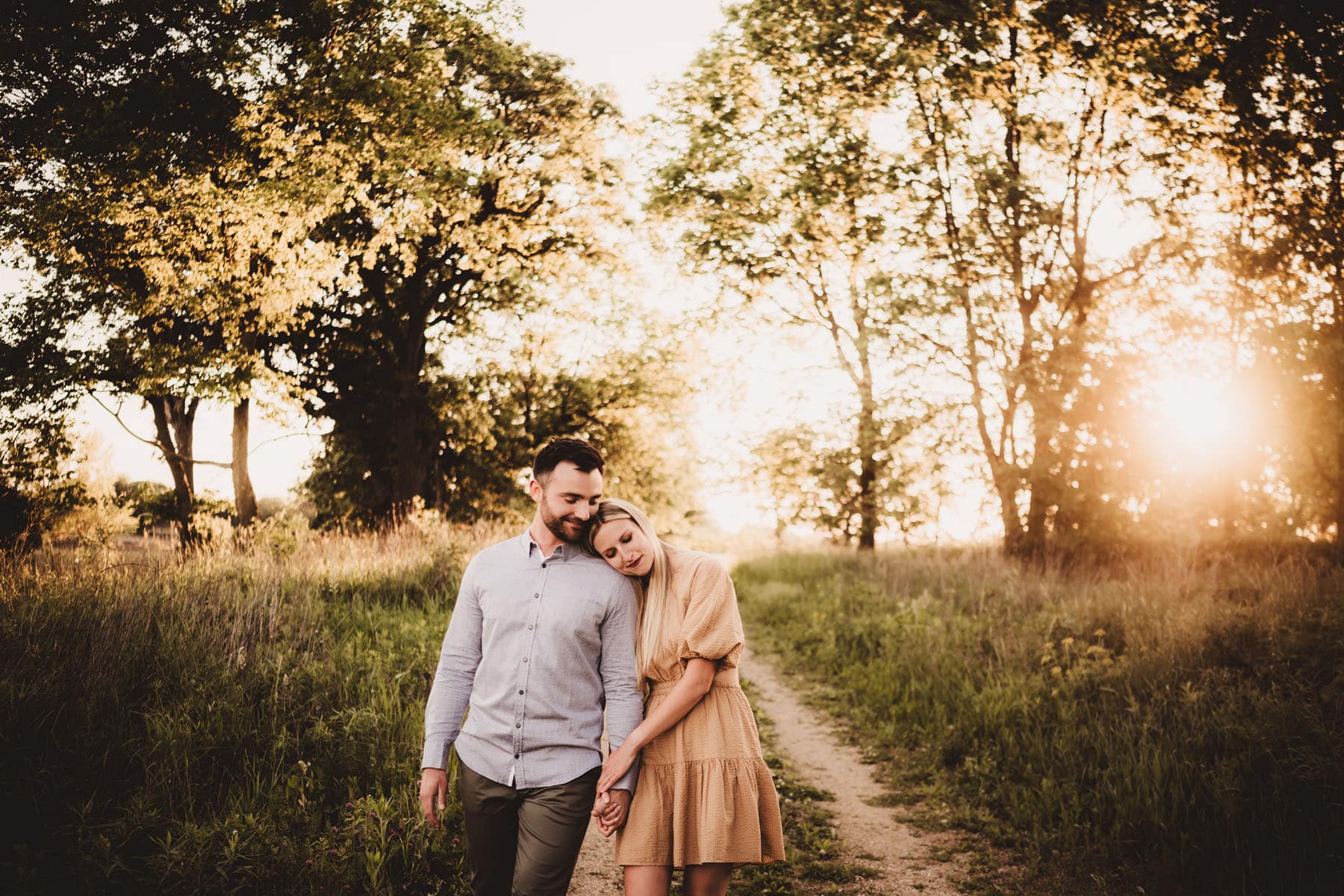 A couple walking hand in hand on a nature path with sun setting through trees in the background
