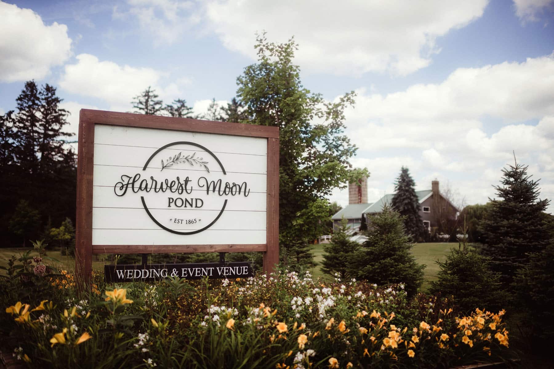 A vintage-style sign for Harvest Moon Pond, a wedding and event venue, surrounded by lush greenery and flowering plants