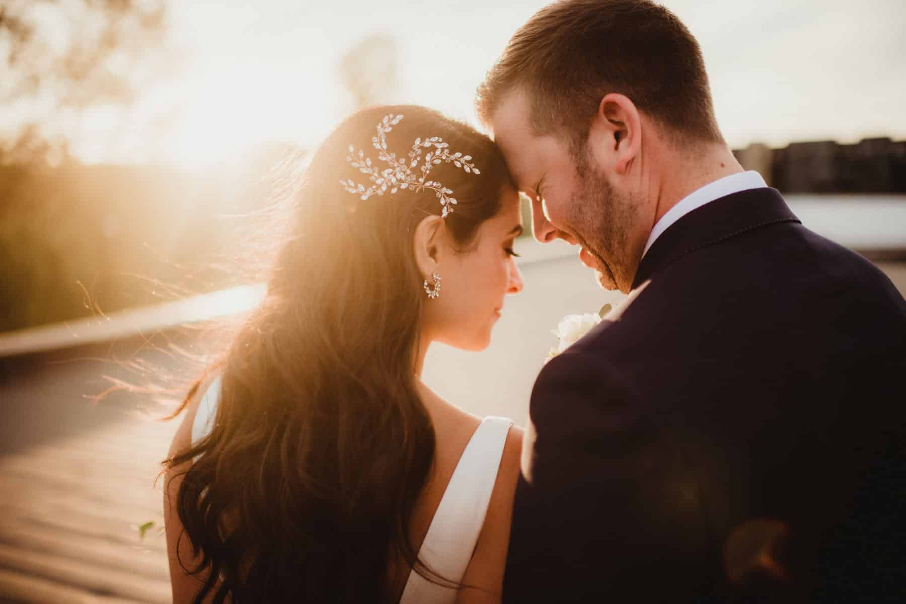 A bride and groom sharing a tender moment with their foreheads touching, backlit by a golden sunset.