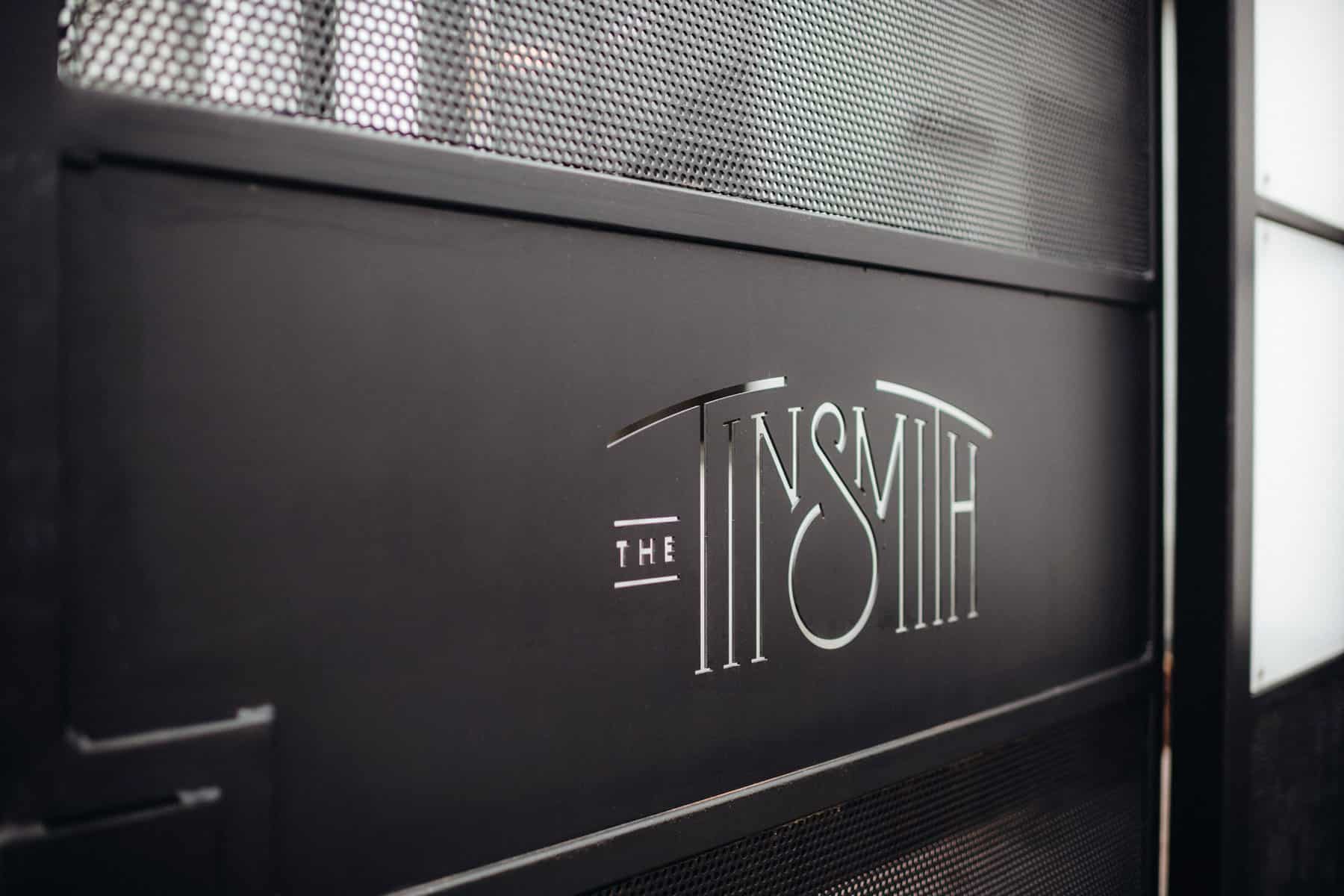 Close-up view of an elegant black metal gate featuring the stylized white logo 'The Tinsmith'.