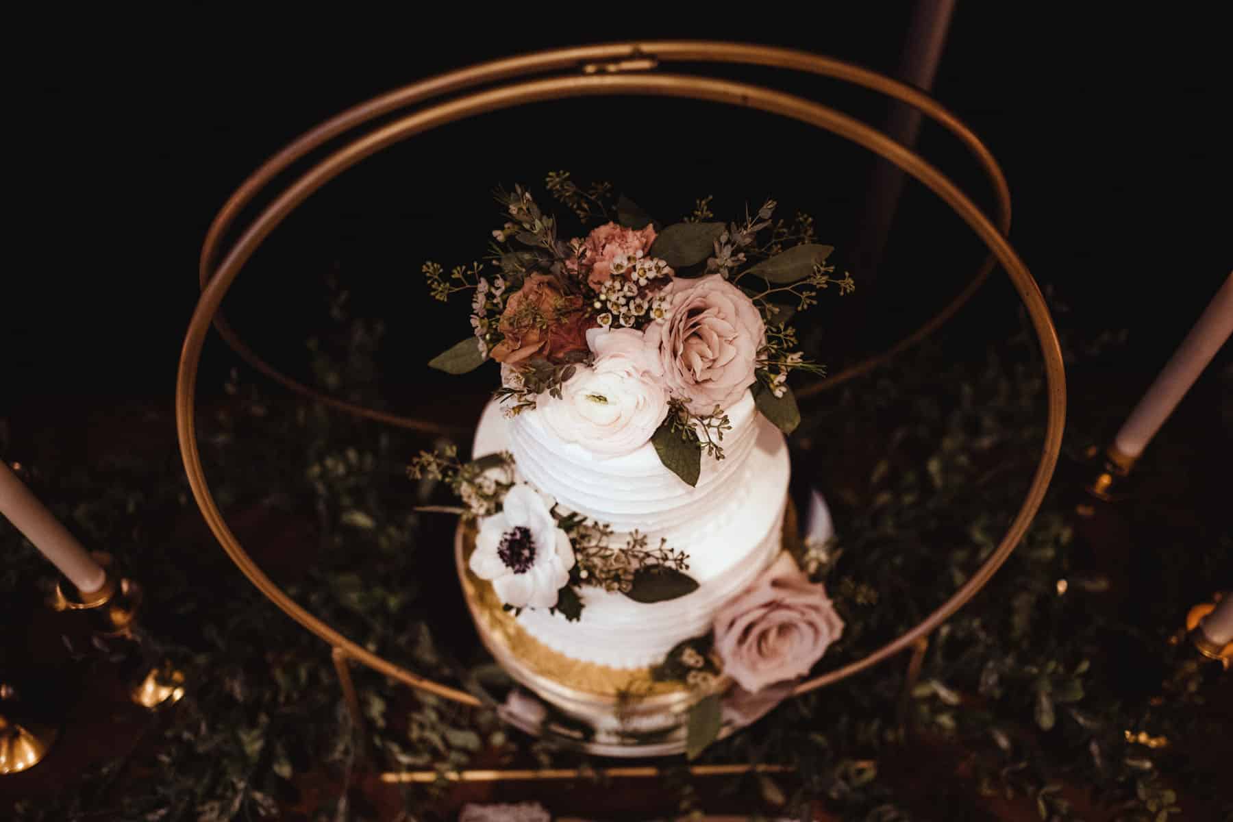 A beautifully decorated wedding cake with layers of white icing and adorned with blush pink roses and assorted greenery, displayed on a golden stand surrounded by foliage in a dimly lit setting.