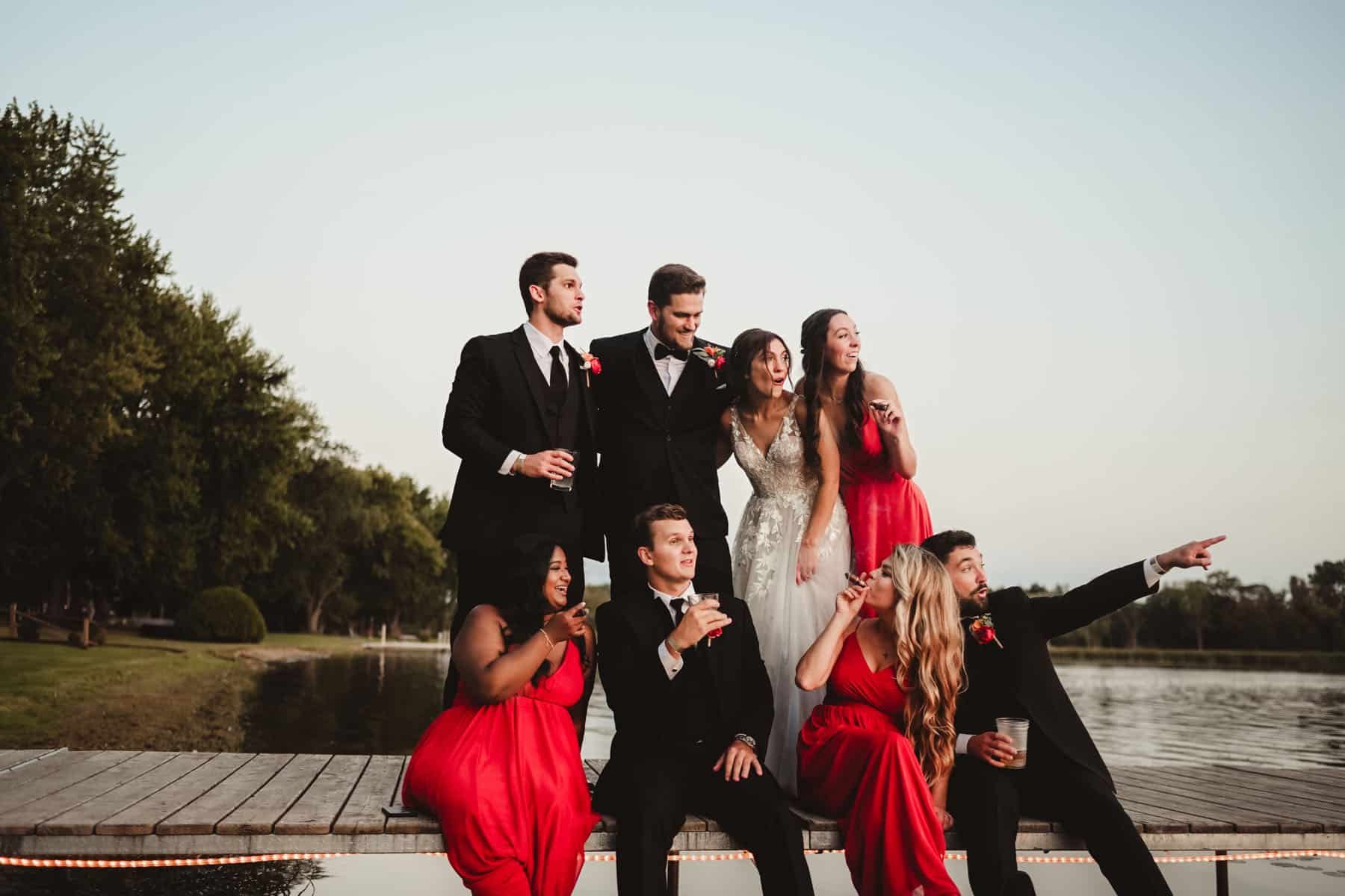 A lively wedding group in elegant attire, with three men and five women, celebrating on a lakeside dock at sunset.