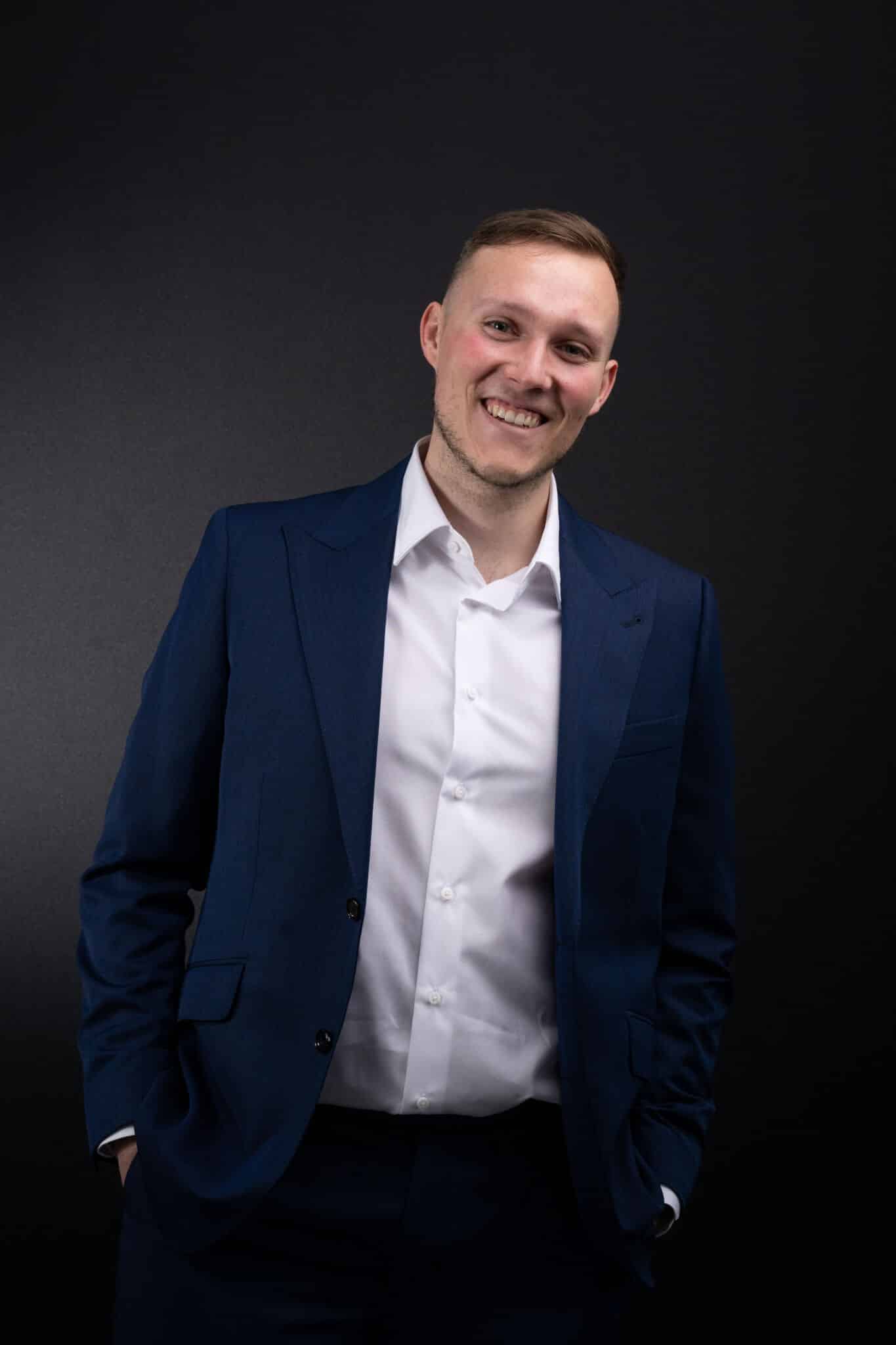 professional_man_in_blue_suit_smiling Confident young man smiling in a blue suit and white shirt, standing against a dark grey background