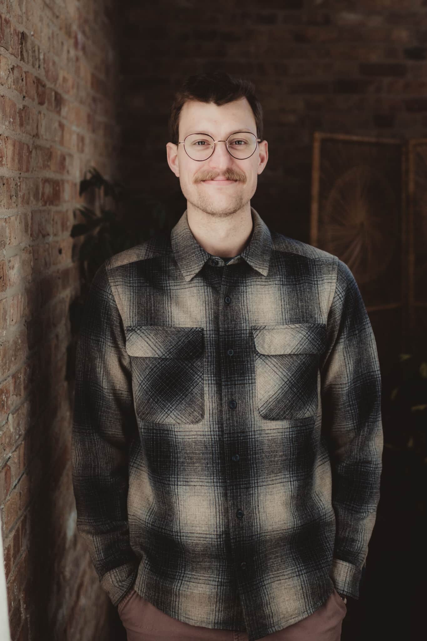 confident_man_with_glasses_indoor_brick_background Smiling man with glasses wearing a checkered shirt standing in front of a brick wall