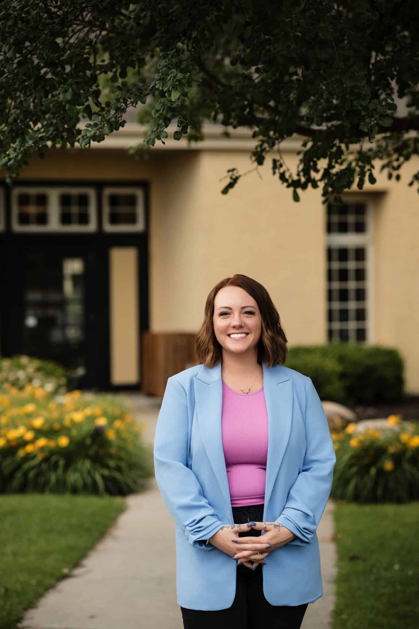 professional_woman_in_blue_blazer_outdoors Confident young woman wearing a blue blazer and pink top, standing with hands clasped in front of a scenic building and green foliage.