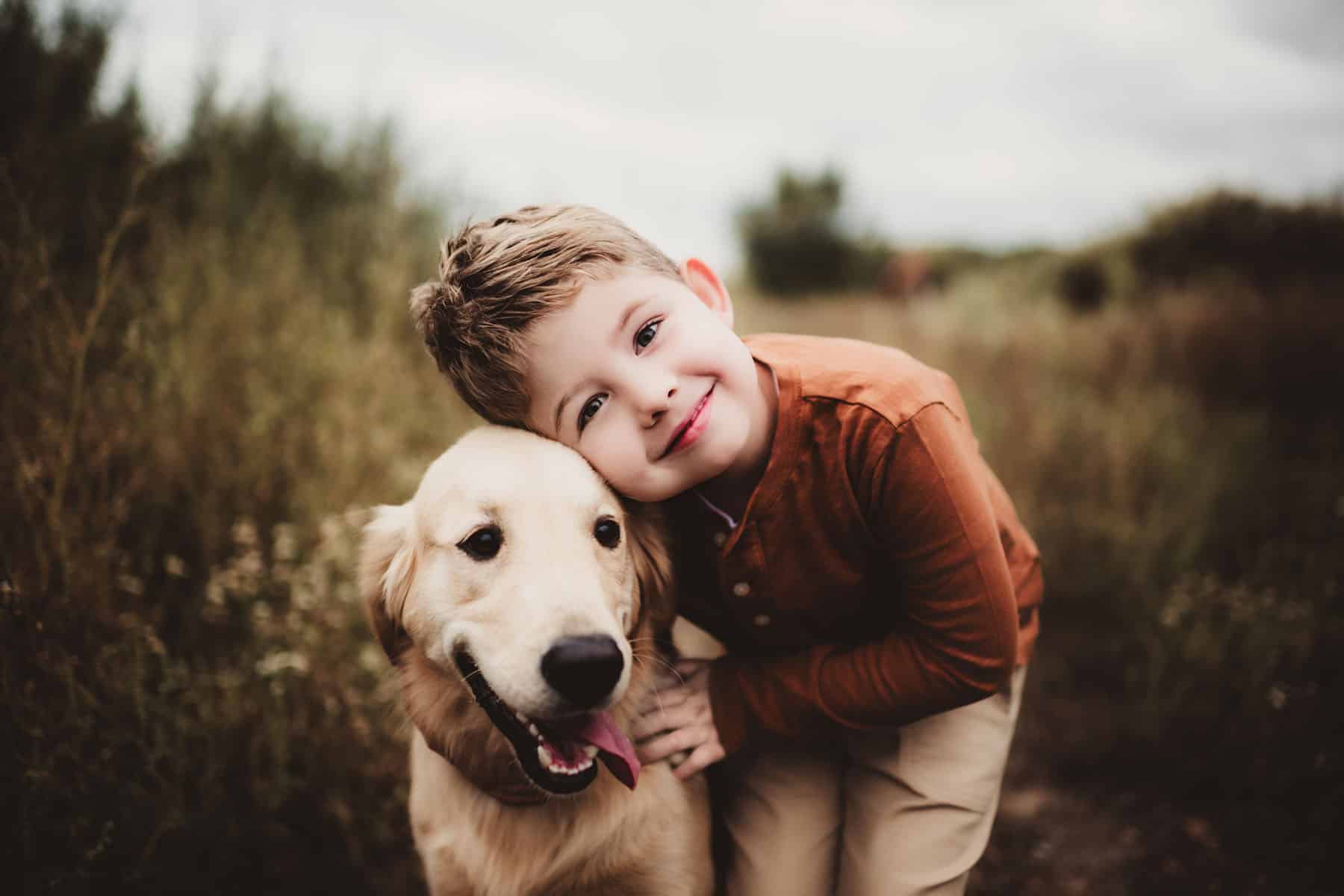 Young boy in a rusty shirt embracing a smiling golden retriever in a natural setting