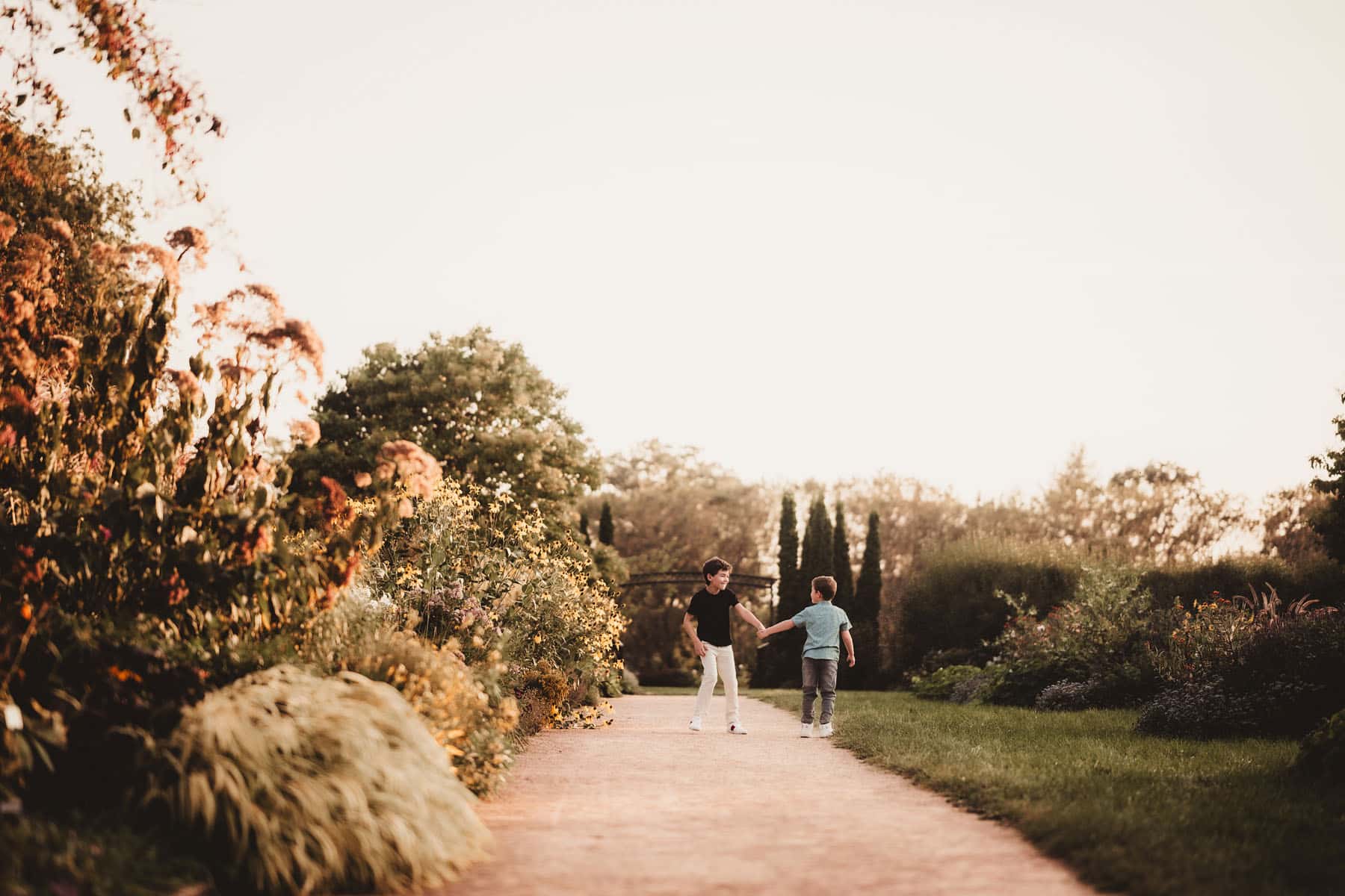 Two young boys walking hand in hand on a garden path surrounded by lush flowers at sunset