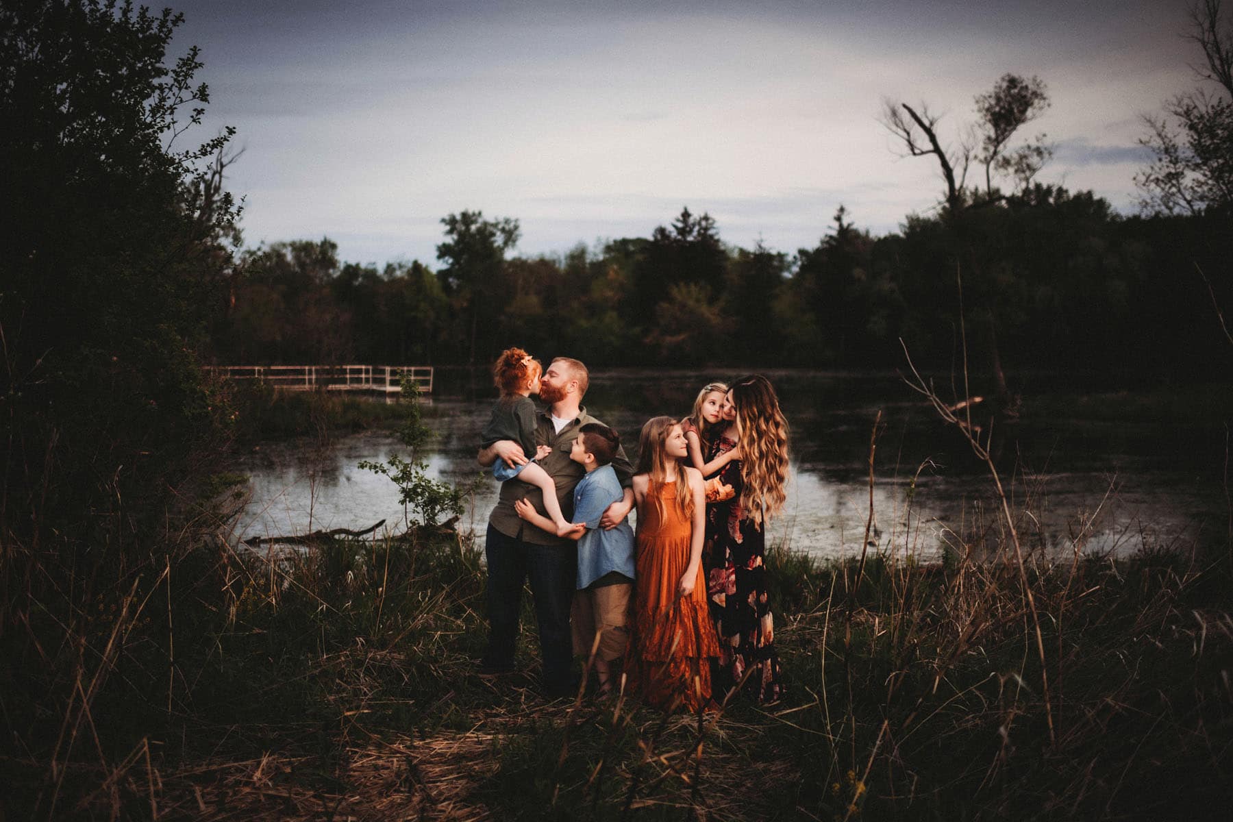 A family of seven with children of various ages embraces lovingly near a serene river at dusk, surrounded by lush vegetation.