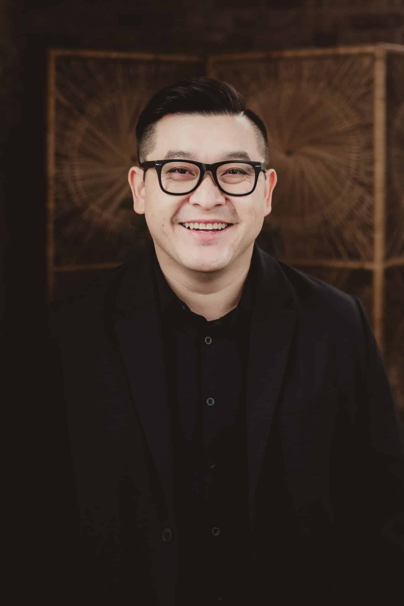 professional_asian_man_in_black_blazer_smiling Confident Asian man wearing black blazer and glasses, smiling in front of a decorative geometric backdrop