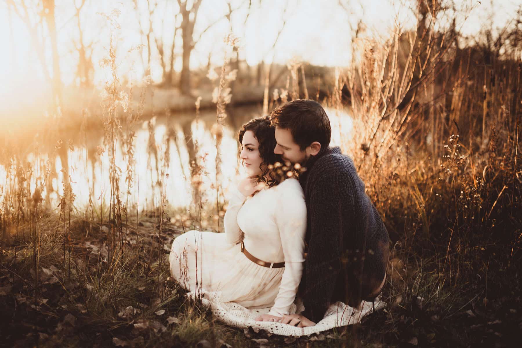 A couple in a loving embrace, seated by a serene lake surrounded by golden sunlight filtering through branches