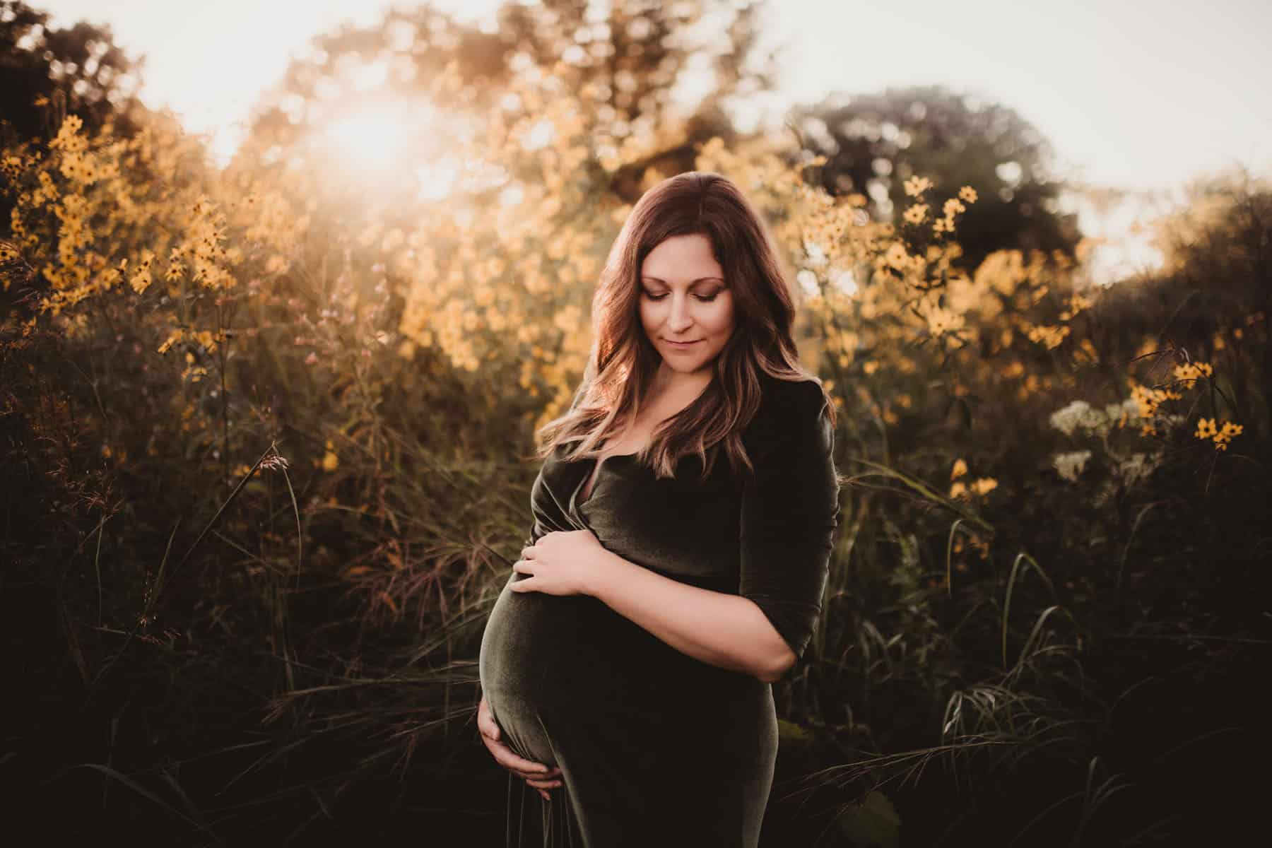 Pregnant woman holding her belly, standing in a field with yellow flowers during sunset