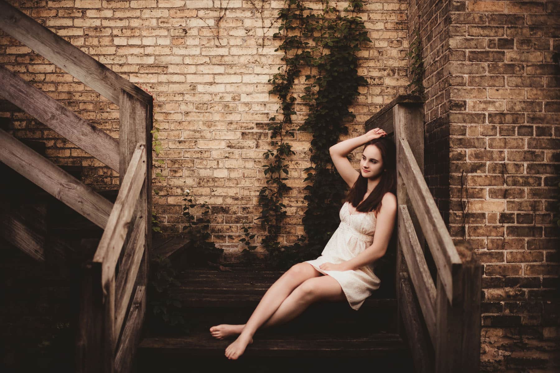 Young woman in a white dress sitting gracefully on wooden stairs against an aged brick wall overgrown with ivy