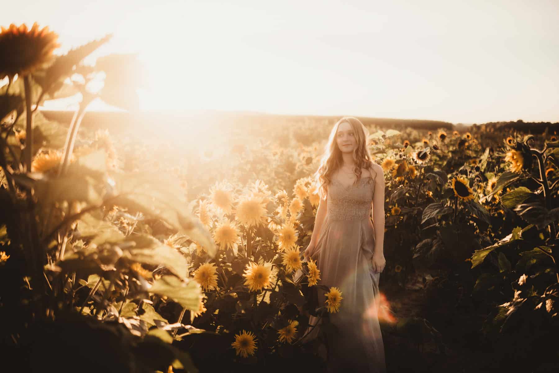 A young woman in a flowing dress walking through a sunlit sunflower field during golden hour