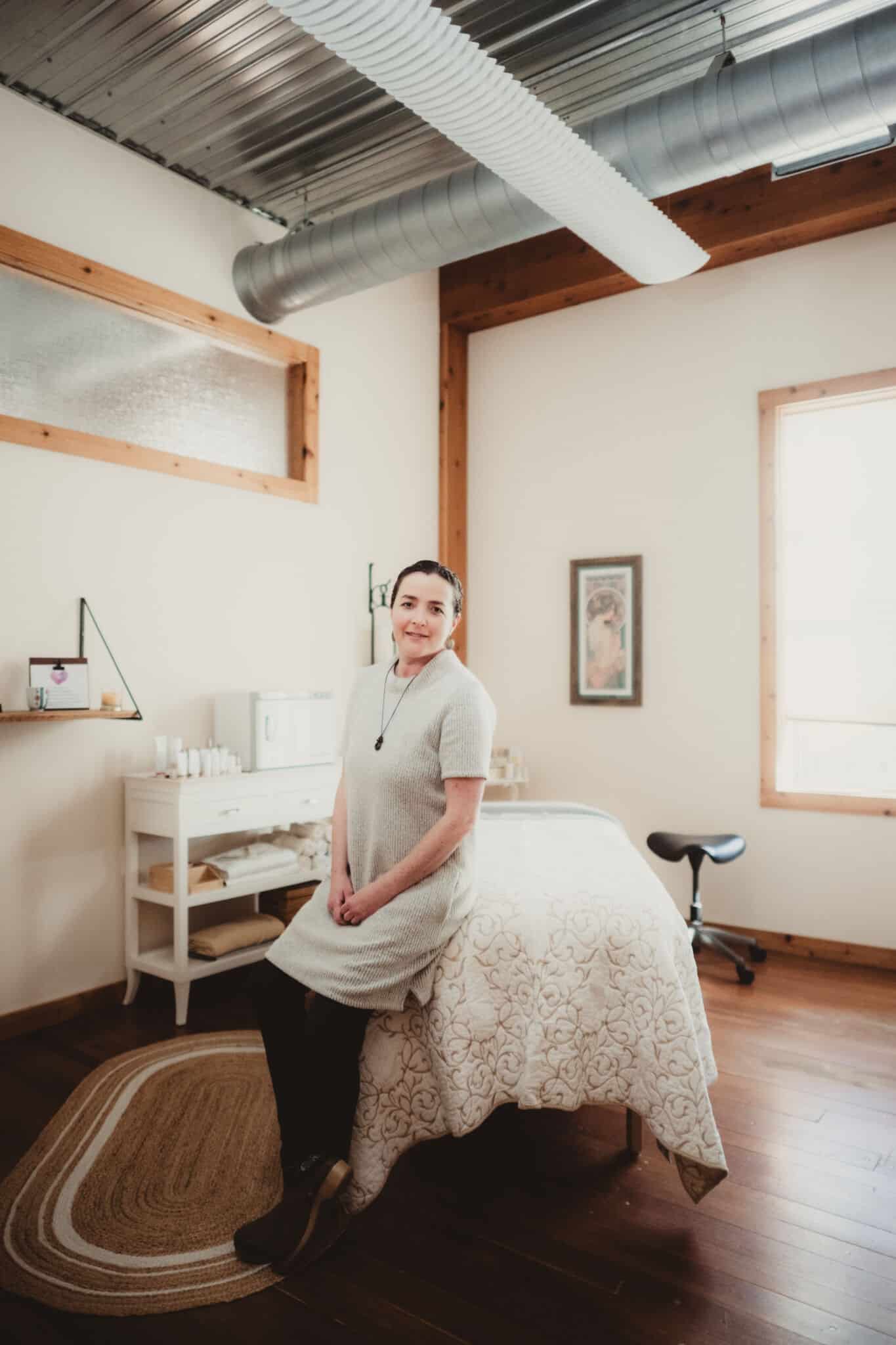 A professional woman standing confidently in a spa room with a massage table, wooden flooring, and minimalist decor.