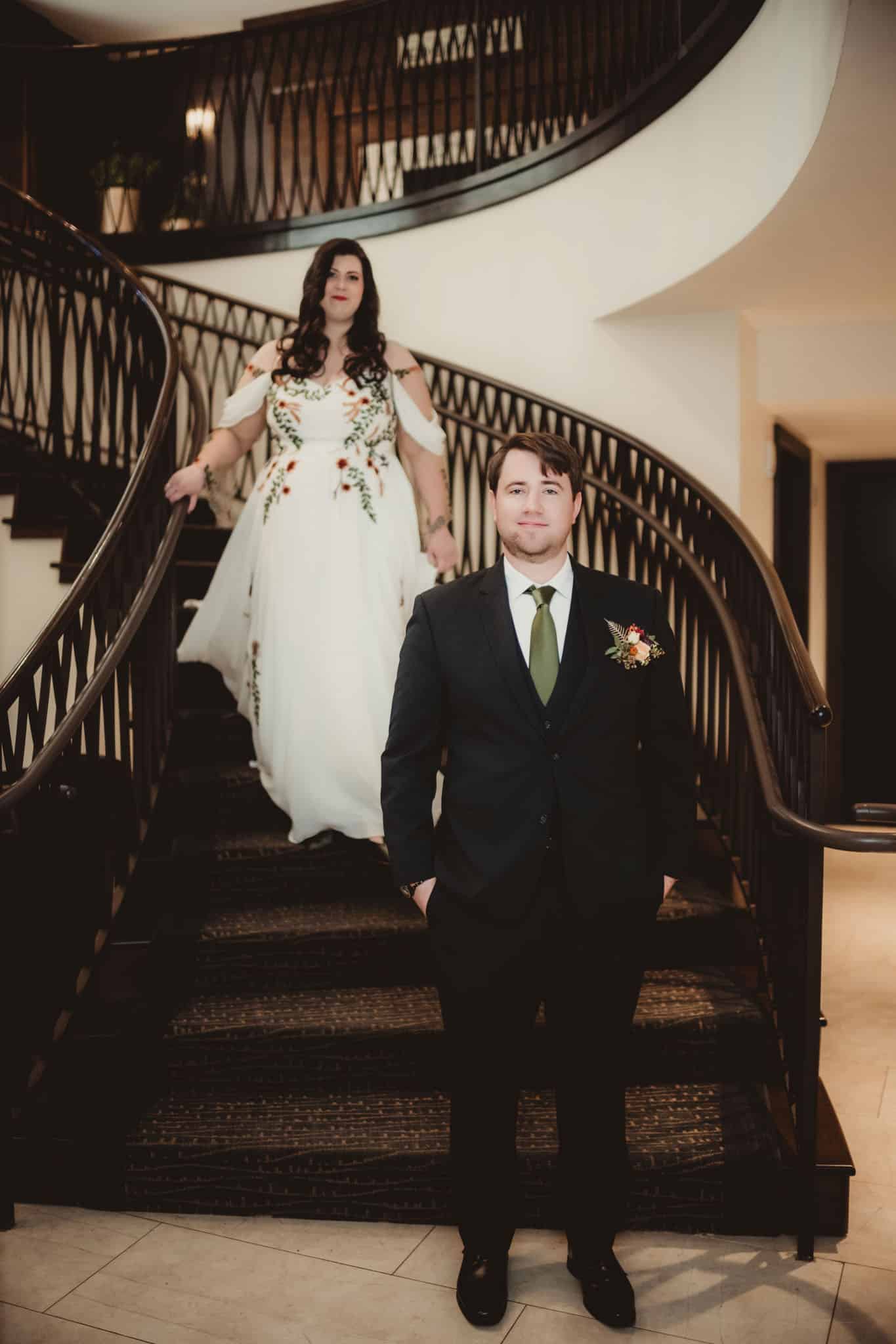 wedding_couple_elegant_staircase A groom in a dark suit and a bride in a flowing white dress with floral embroidery descending a grand spiral staircase.