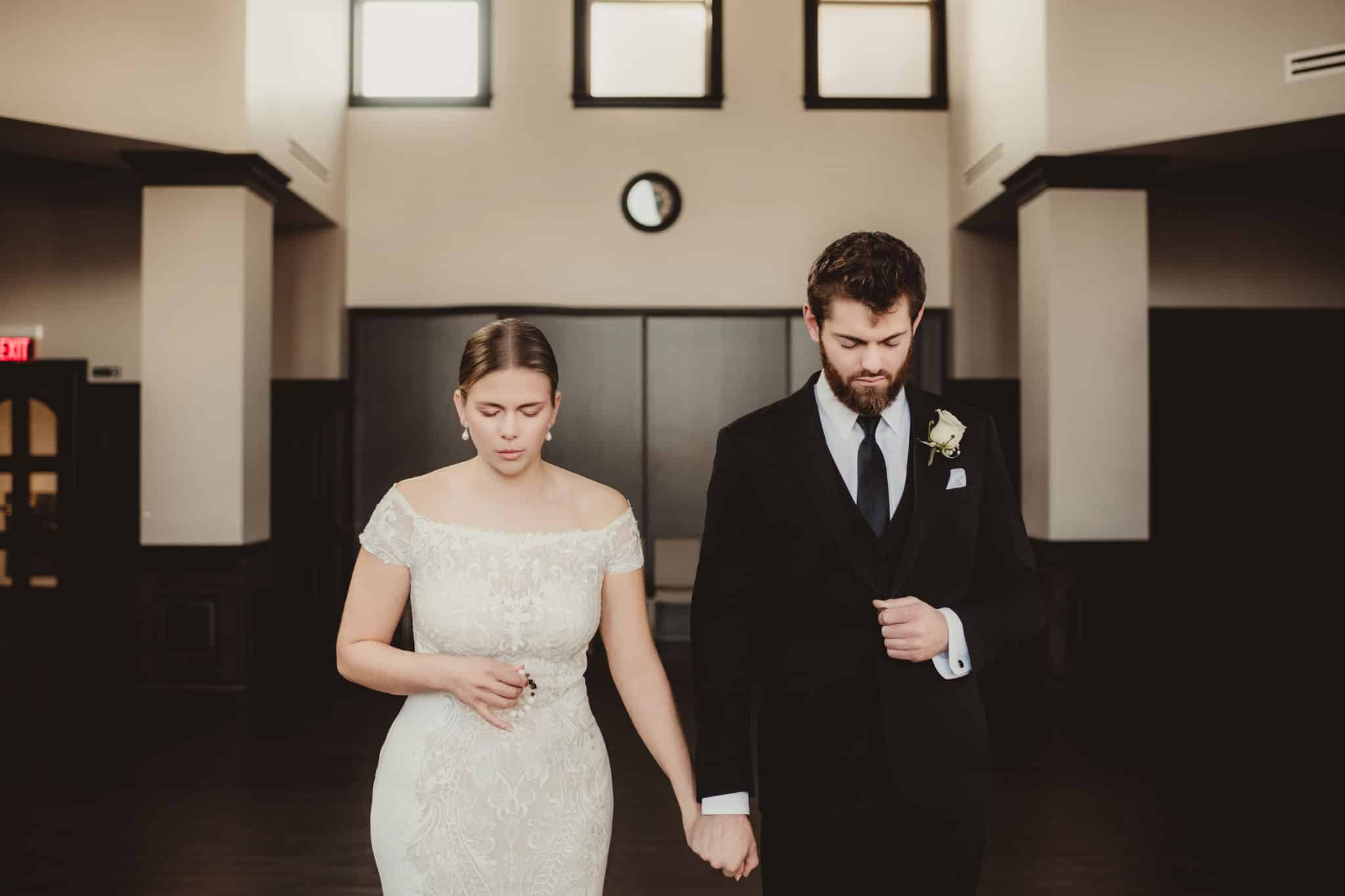 elegant_wedding_couple_walking_hand_in_hand A bride in a laced wedding dress and a groom in a black suit walking hand in hand, looking contemplative