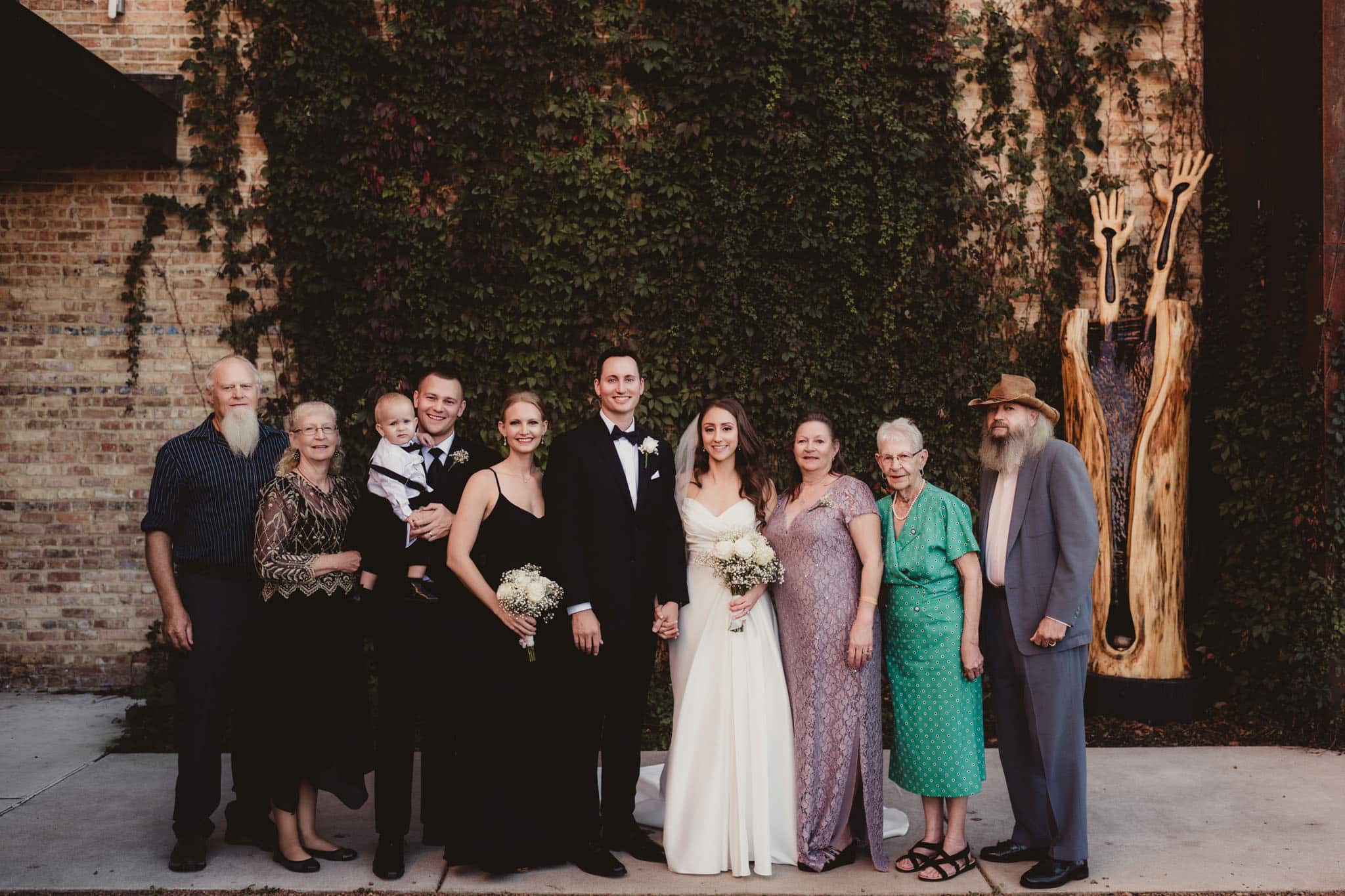 multi_generational_family_wedding_outdoors_vintage_brick_wall A multigenerational family dressed in formal wear standing together in front of an ivy-covered brick wall at a wedding